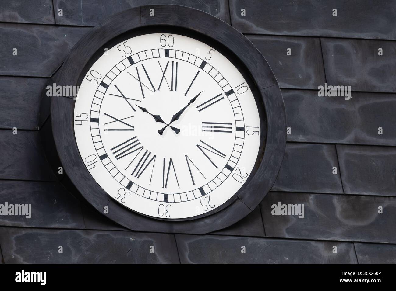 Eine auffällige große Runduhr auf einem dunklen Dach mit kräftigen römischen Ziffern und einem weißen Gesicht. Eleganter Zeitmesser, ideal für Vintage, Architektur, Stockfoto
