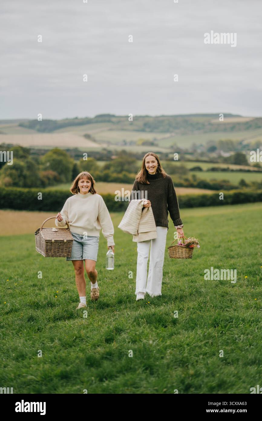 Zwei junge Frauen schlendern durch ein üppiges grünes Feld, von denen eine einen Korb trägt. Die Szene ist ruhig und ruhig, die Frauen genießen das B Stockfoto