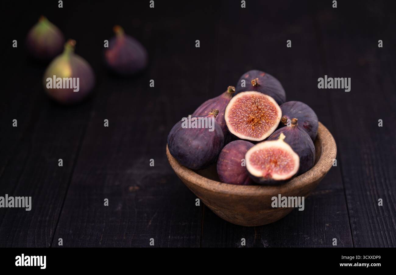 Zwei in Scheiben geschnittene Feigen in einer Holzschale neben ganzen Feigen auf dunklem Holztisch, stimmungsvolles Stillleben Stockfoto
