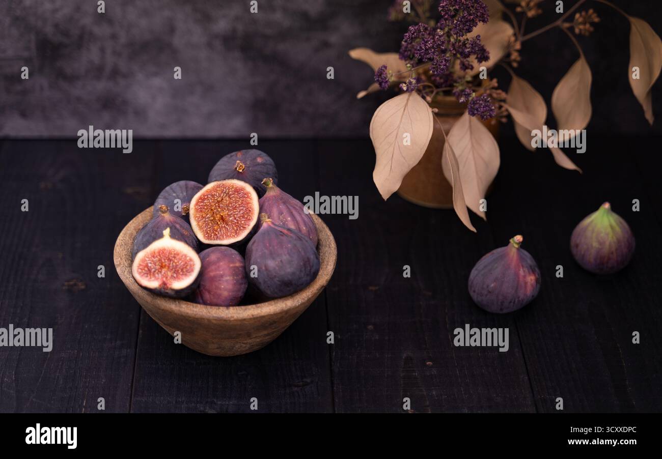 Zwei in Scheiben geschnittene Feigen in einer Holzschale neben ganzen Feigen auf dunklem Holztisch, stimmungsvolles Stillleben mit Vasendekor hinten Stockfoto