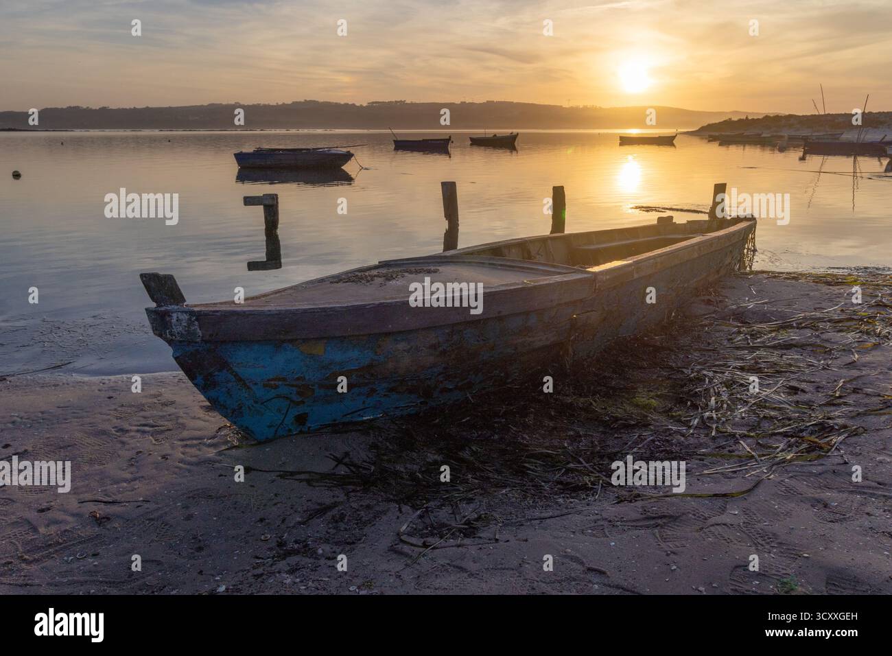 Altes Boot ruht an einem Sandstrand während eines ruhigen Sonnenaufgangs mit Reflexionen auf dem Wasser Stockfoto