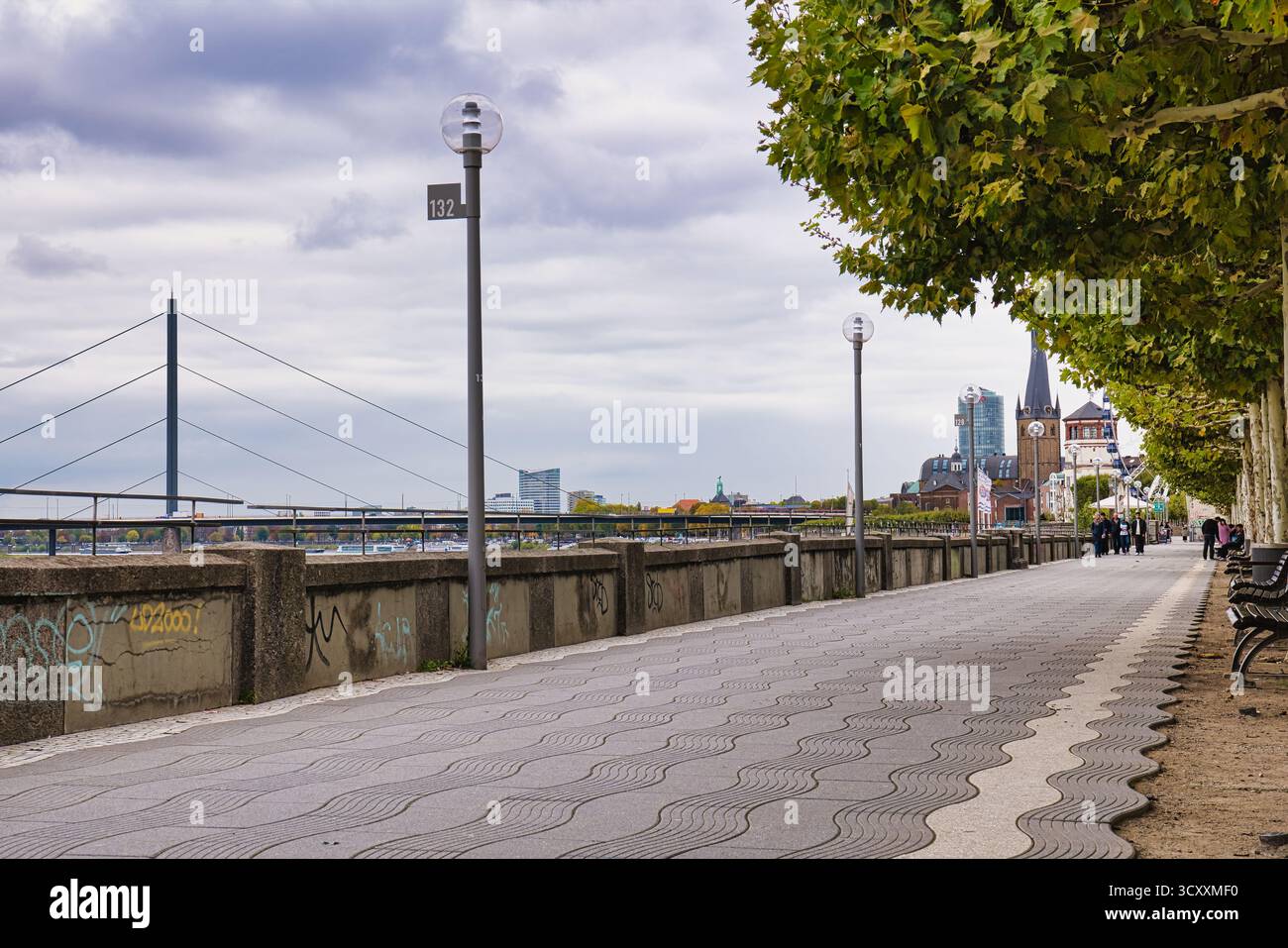 Leere, von Bäumen gesäumte Promenade entlang des Rheins mit weit entferntem Kirchturm und urbaner Skyline im bewölkten Herbst Düsseldorf Deutschland Stockfoto