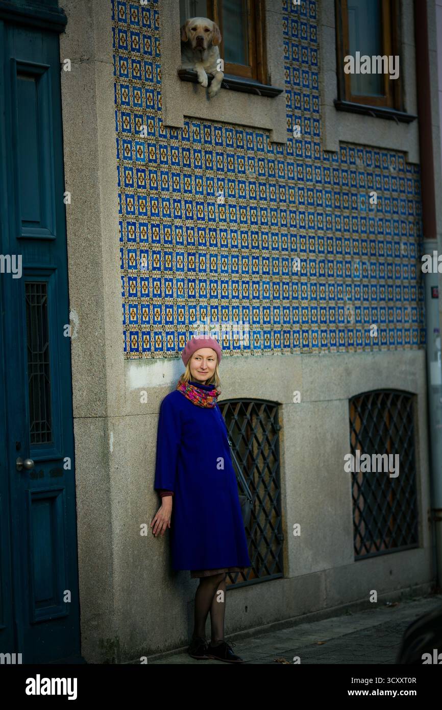 Eine Frau in Fliedermantel und Baskenmütze steht an einer mit Azulejos bedeckten Wand in Porto, Portugal, während ein Hund vom Fenster hinunter blickt. Die Szene f Stockfoto