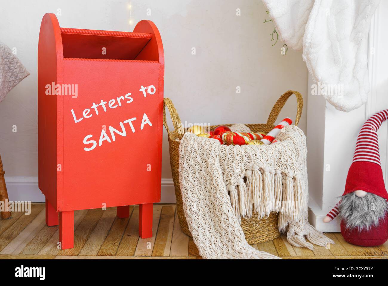 Festliche Weihnachtsszene mit rotem Briefkasten für Weihnachtsbriefe, Korb mit Ornamenten, Strickdecke, skandinavischem Zwerg oder Elfe auf Holzboden. Winter Stockfoto