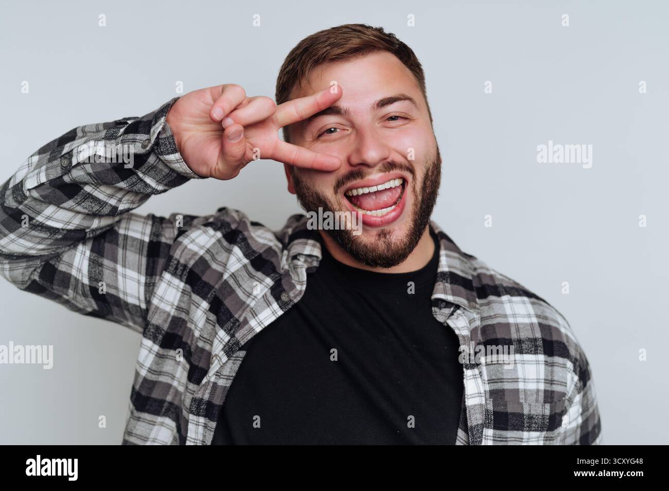Ein Mann lächelt breit, während er mit der Hand ein Friedenszeichen macht. Er trägt ein kariertes Hemd über einem schwarzen T-Shirt und ist in einem hellen, neutralen Setting. Sein Stockfoto