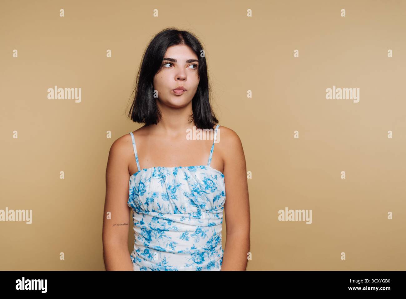 Eine junge Frau steht in einem Studio und trägt ein Blumenoberteil. Sie schaut mit einem nachdenklichen Ausdruck auf ihrem Gesicht weg. Der neutrale Hintergrund fügt den Fokus hinzu Stockfoto