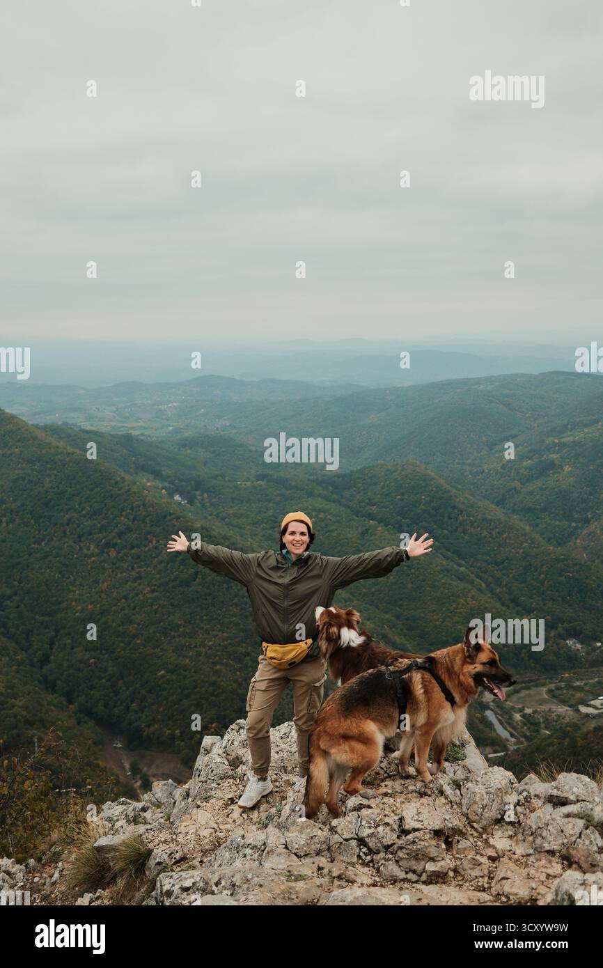 Eine glückliche Frau hebt ihre Arme auf dem Gipfel des Berges Kablar, Serbien, mit ihren beiden Hunden Australian und German Shepherd neben ihr, und genießt die Freiheit Stockfoto