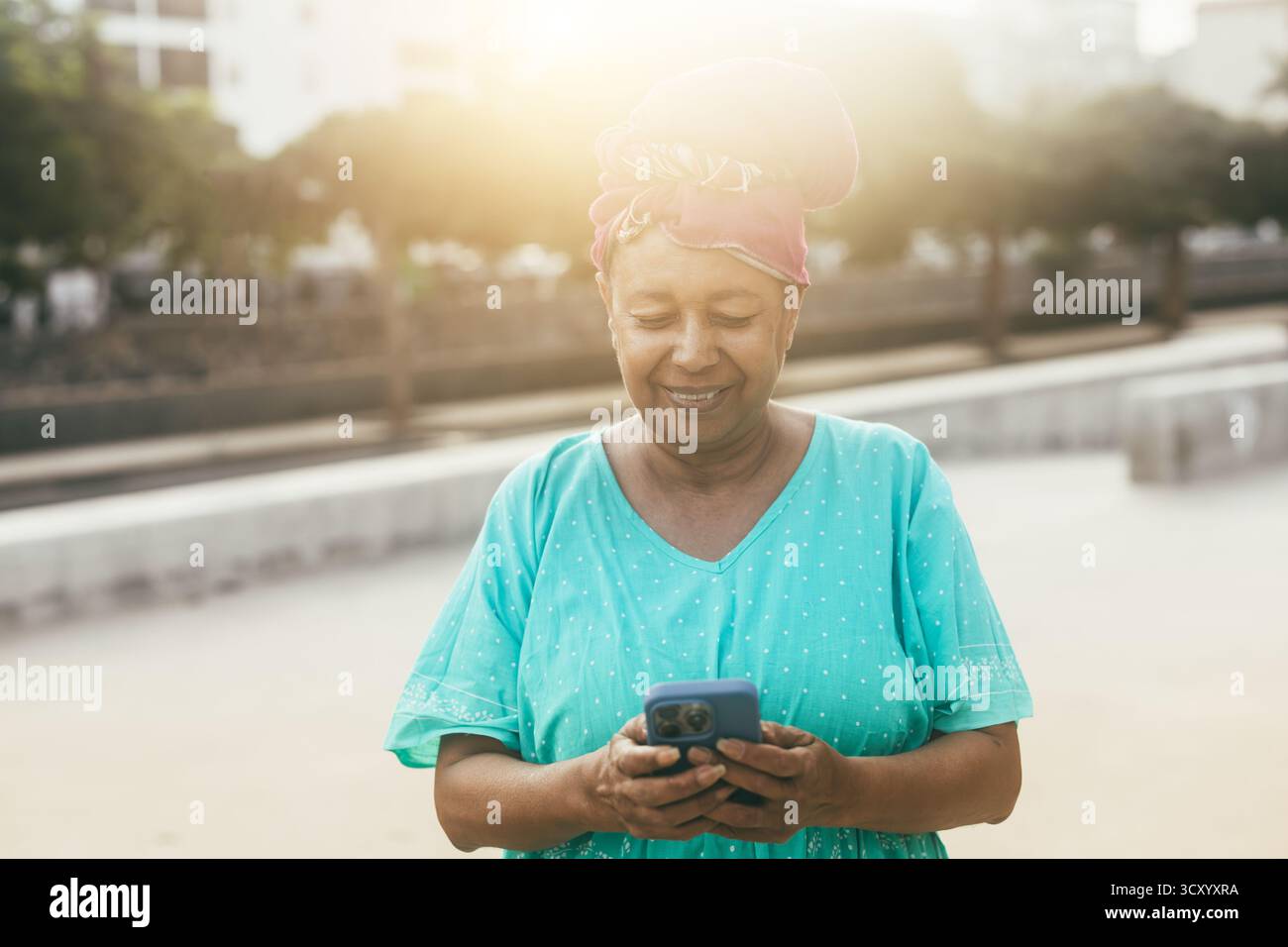 Glückliche afrikanische Seniorin mit Smartphone im Freien mit Stadt im Hintergrund, die traditionelle Kleidung trägt - fröhlicher älterer Lebensstil und Technologie Stockfoto