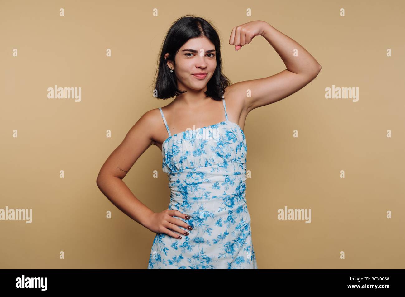 Eine junge Frau beugt ihren Arm selbstbewusst und trägt ein blaues Blumenkleid. Sie steht vor einem hellbraunen Hintergrund und zeigt ihre Stärke und Begeisterung Stockfoto