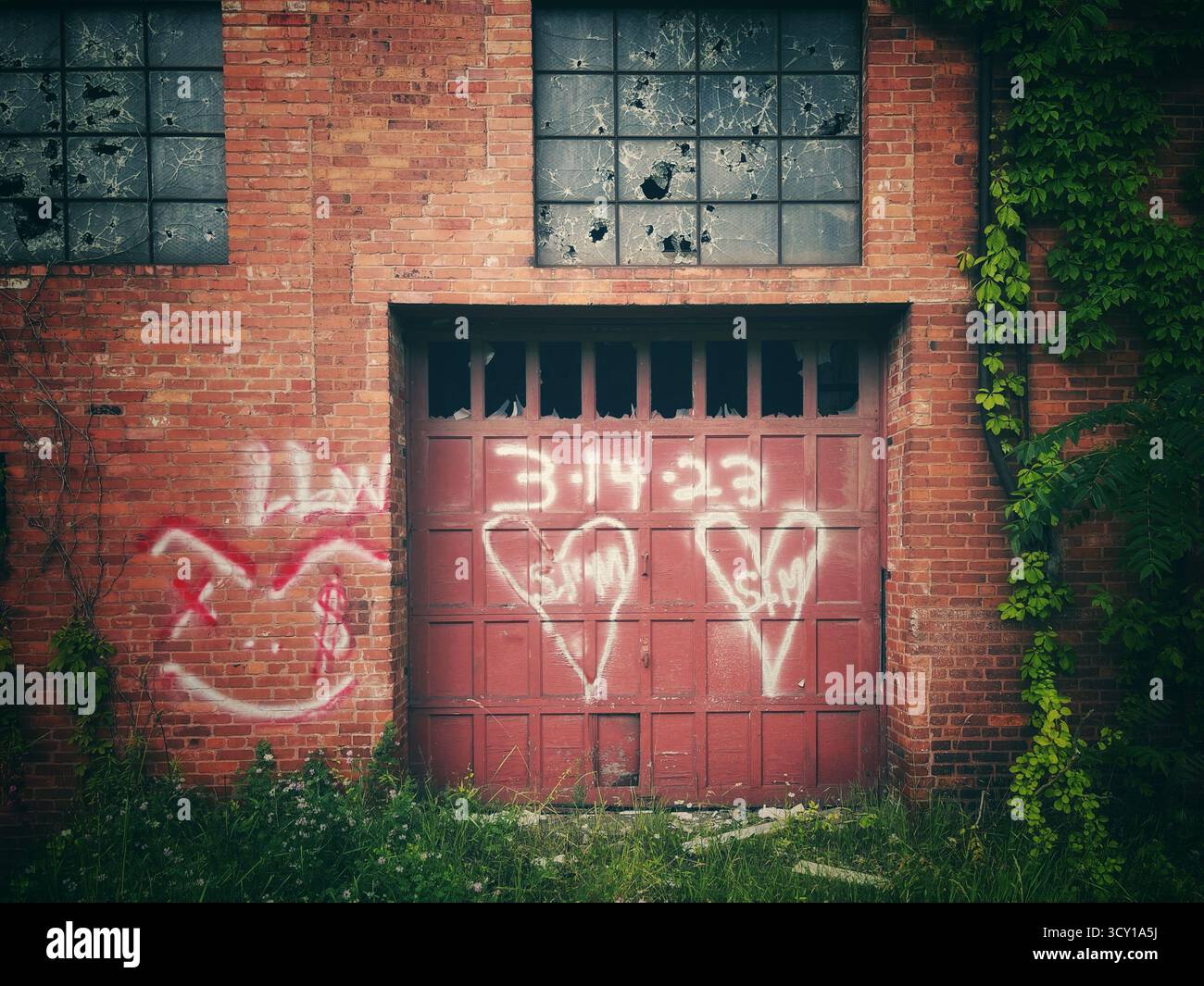 Altes industrielles Backsteingebäude mit rotem Garagentor, bedeckt mit Graffiti und umgeben von bewachsenen Pflanzen und Weinstöcken Stockfoto