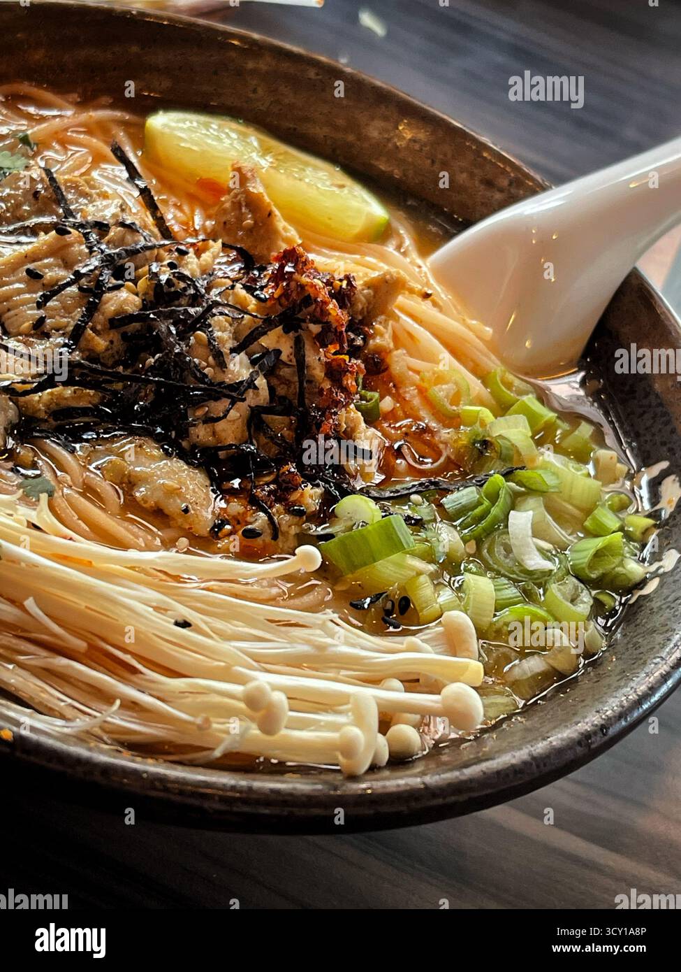 Nahaufnahme einer dampfenden Schüssel Ramen mit Enoki-Pilzen, grünen Zwiebeln, Algen und einer Scheibe Zitrone darauf - Smartphone-aufgenommenes Stockfoto