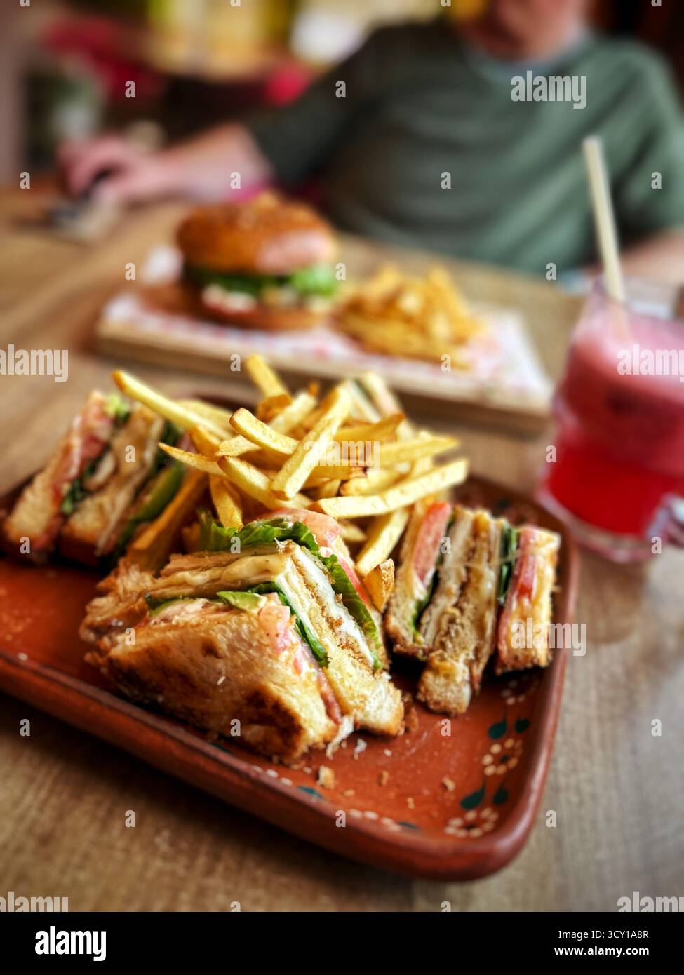 Nahaufnahme eines köstlichen Club-Sandwiches mit knusprigen Pommes, serviert auf einem Teller, mit einem Burger und Getränk im Hintergrund auf einem Holztisch Stockfoto