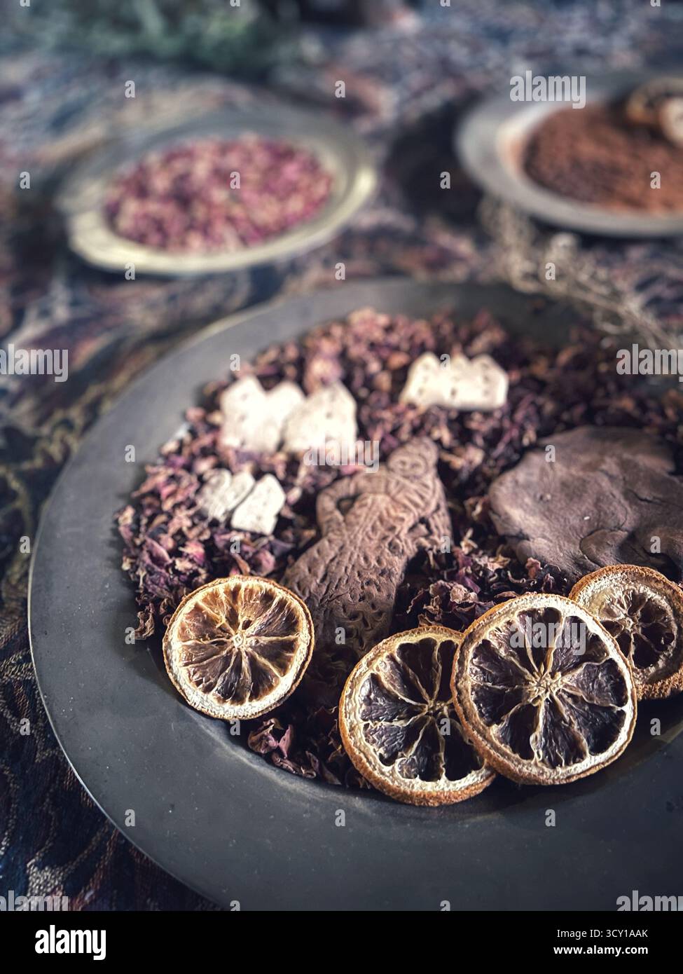 Getrocknete Zitrusscheiben und verschiedene Kräuter in einer dekorativen Schüssel schaffen eine elegante Präsentation auf einer gemusterten Tischdecke - Smartphone-aufgenommenes Stockfoto