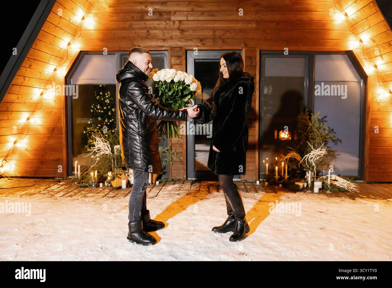 Paare umarmen sich mit einem Blumenstrauß aus weißen Rosen vor einem hölzernen Chalet, das in einer Winternacht mit Lichtern dekoriert ist Stockfoto
