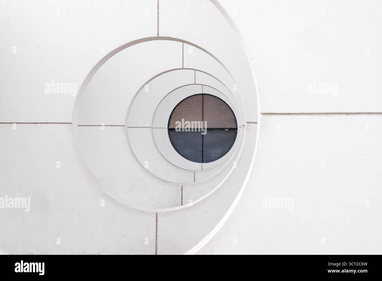 Ein markantes minimalistisches architektonisches Detail mit einem weißen konzentrischen Spiraldesign, das zu einem runden Fenster mit einem dunklen metallischen Gitter führt. Stockfoto