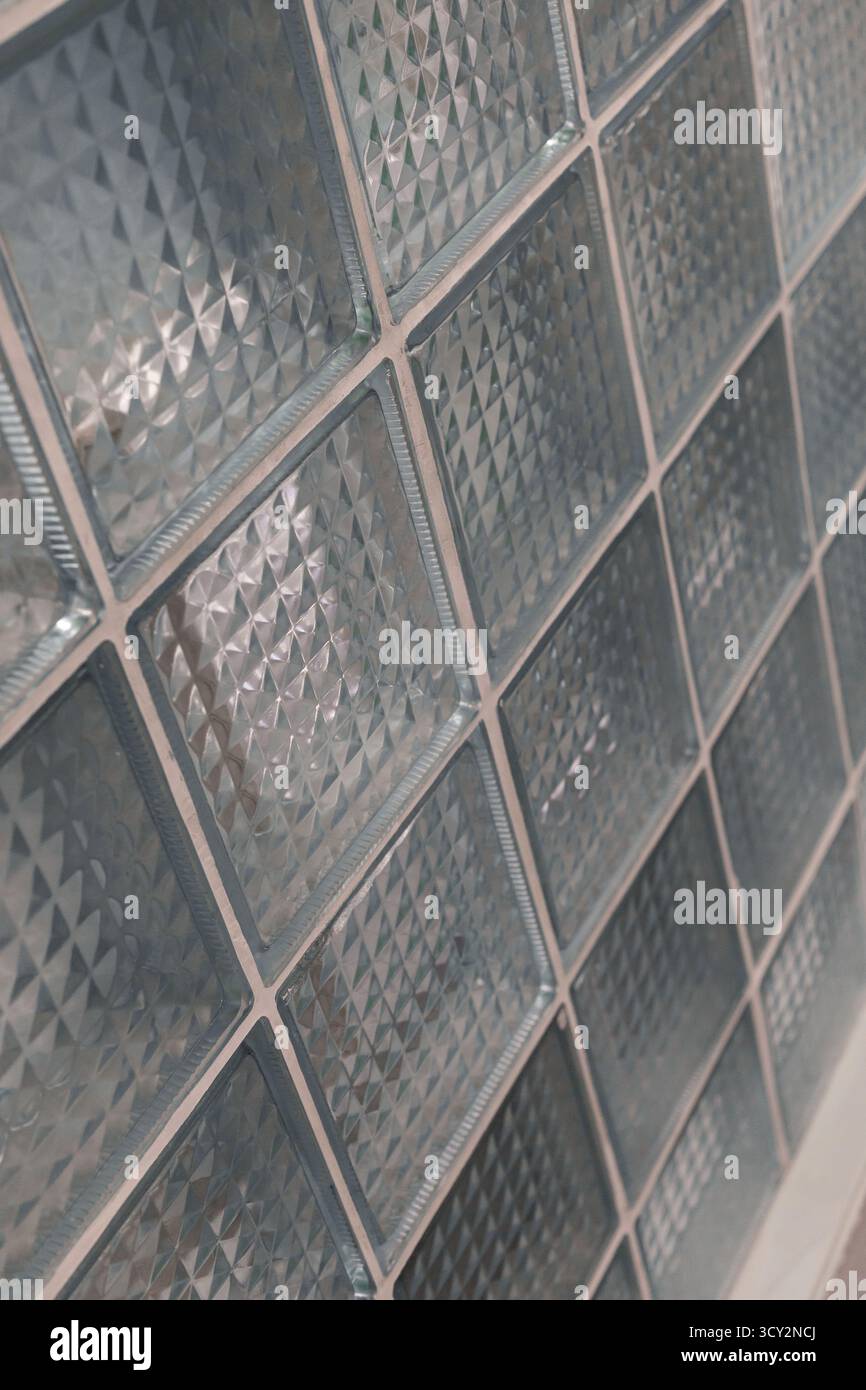 Eine Wand aus klaren Glasblöcken mit geometrischen Mustern reflektiert das Licht und verleiht Textur und unterstreicht mit ihrem einzigartigen Design einen modernen Raum. Stockfoto