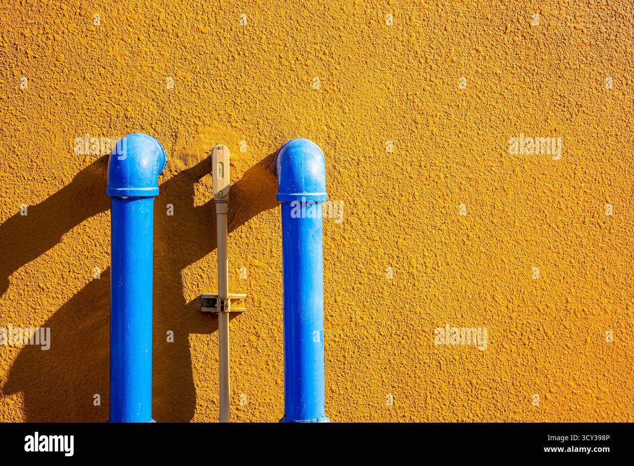 Blaue Wasserrohre auf gelb strukturierter Wand mit Sonnenlicht-Schatten, minimaler industrieller Hintergrund, farbenfroher Kontrast für Architekturdesign und kreatives C Stockfoto