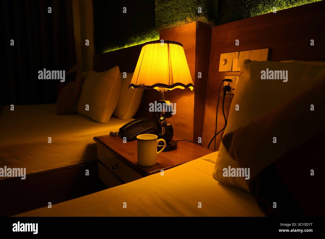 Ein dunkler, gemütlicher Nachttisch im Hotelzimmer mit einer gelb leuchtenden Tischlampe, einem Telefon und einem weißen Becher, der das warme, einladende Ambiente zwischen zwei und zwei unterstreicht Stockfoto