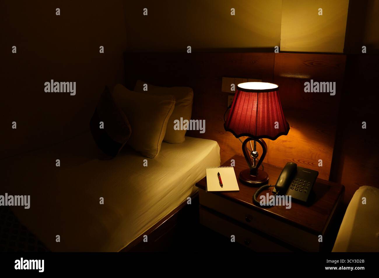 Ein schwach beleuchteter Nachttisch im Hotelzimmer zwischen Bett und Wand, mit einer leuchtend roten Tischlampe, einem Notizblock mit Stift und einem Telefon. Stockfoto