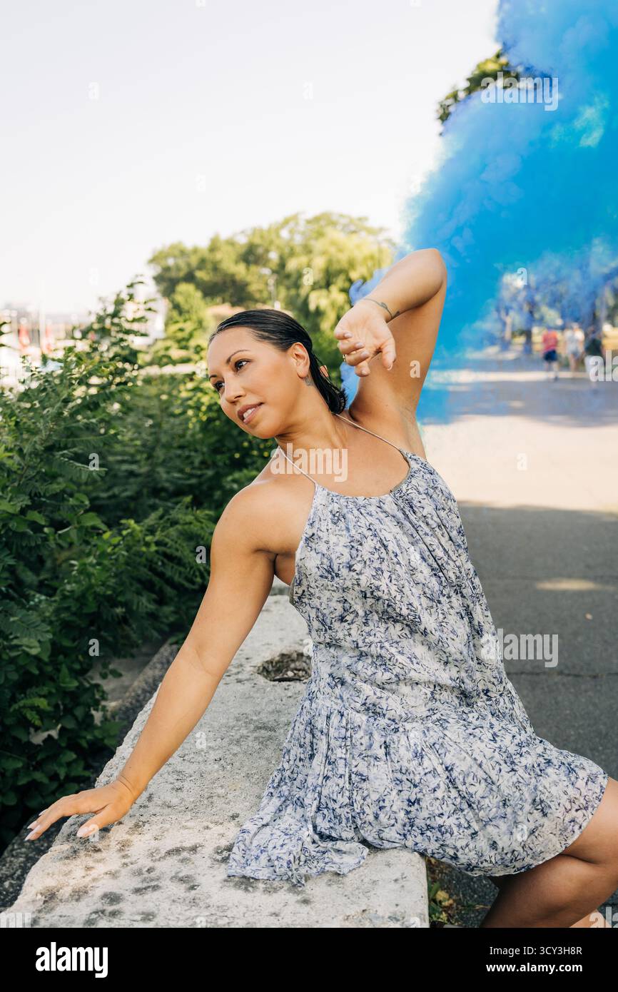 Frau im Blumenkleid posiert mit blauem Rauch im sonnigen Park Stockfoto