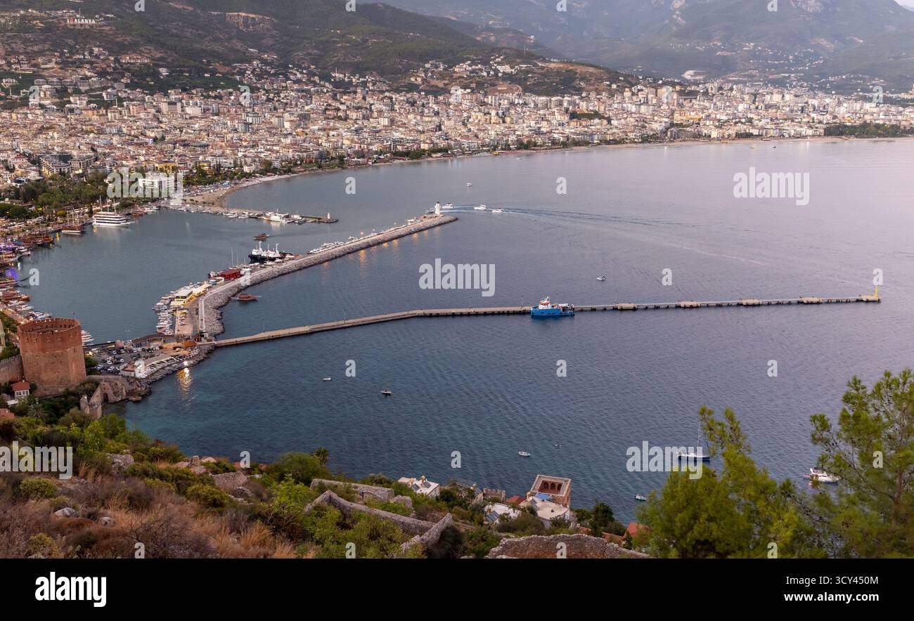 Alanya, Türkei. Panoramablick auf den Hafen mit einem geschwungenen Wellenbrecher, einem blauen Schiff und geschäftigen Piers entlang einer ruhigen Bucht. Berge erheben sich hinter einer dichten Küste Stockfoto