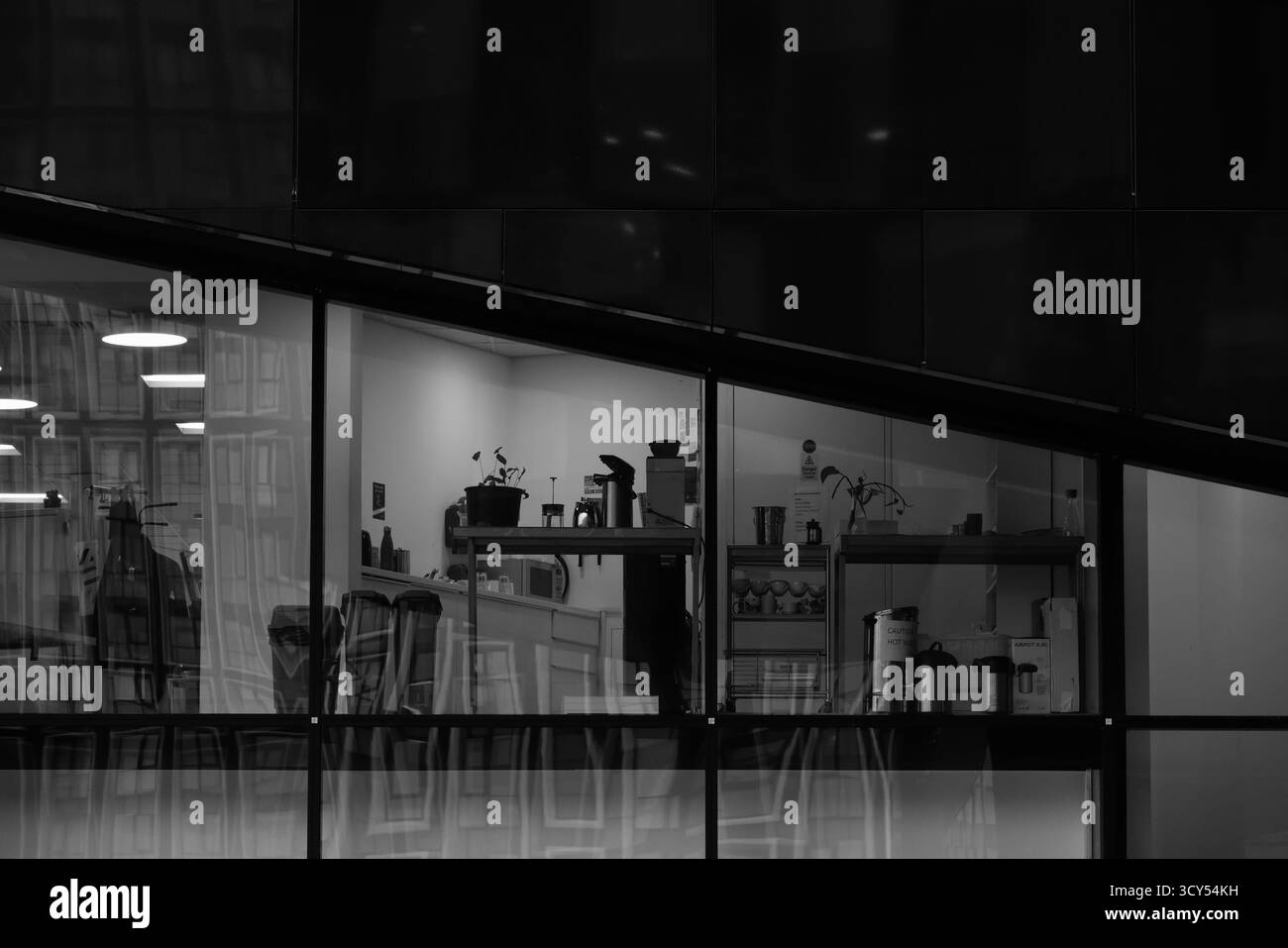 Schwarzweiß-Foto einer modernen Büroküche, die durch Glasfenster gesehen wird, mit Pflanzen, Regalen, Kaffeemaschinen und Reflexen des nahegelegenen Gebäudes Stockfoto