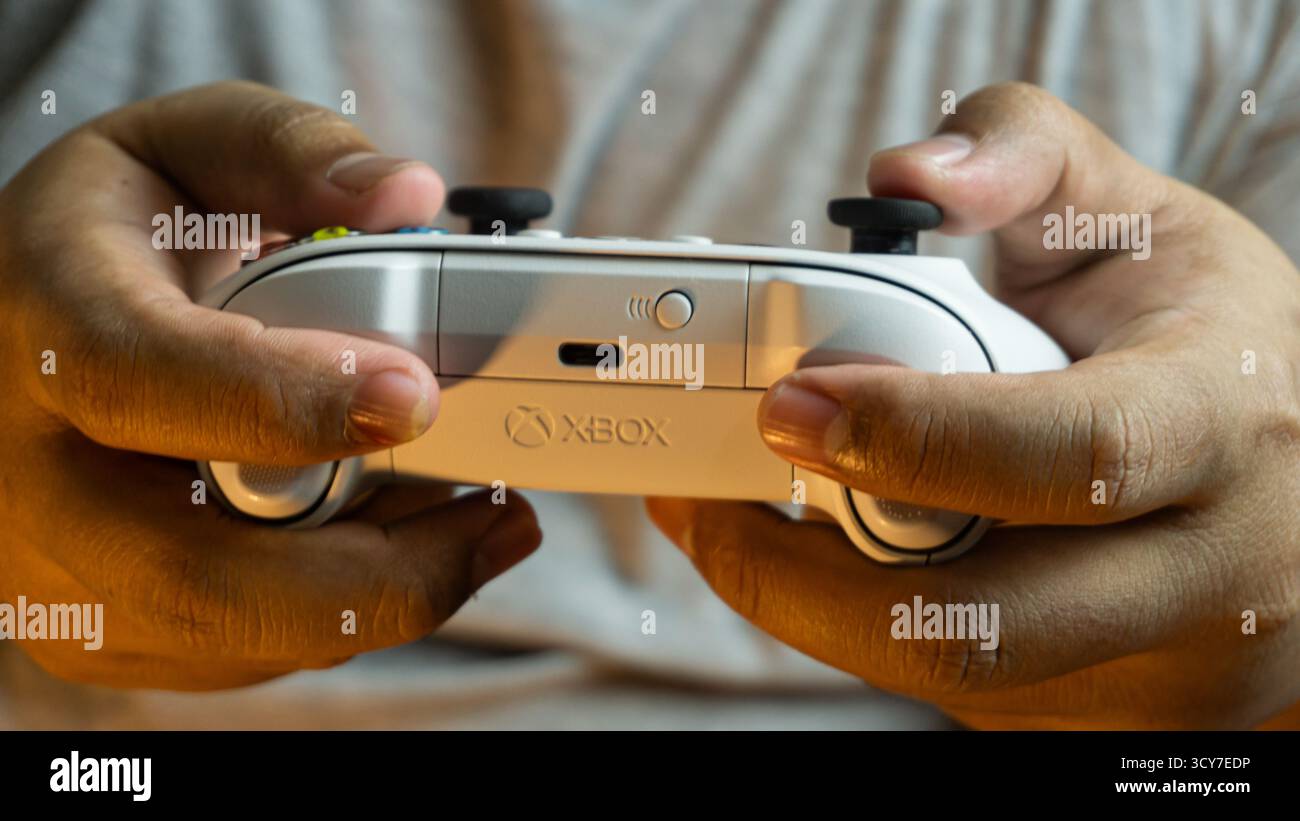 Süd-Kalimantan, Indonesien - 07 14 2025 : Weißer S|X-Controller der Xbox-Serie, gehalten von zwei Händen, mit warmer Umgebungsbeleuchtung, die den komfortablen gri hervorhebt Stockfoto