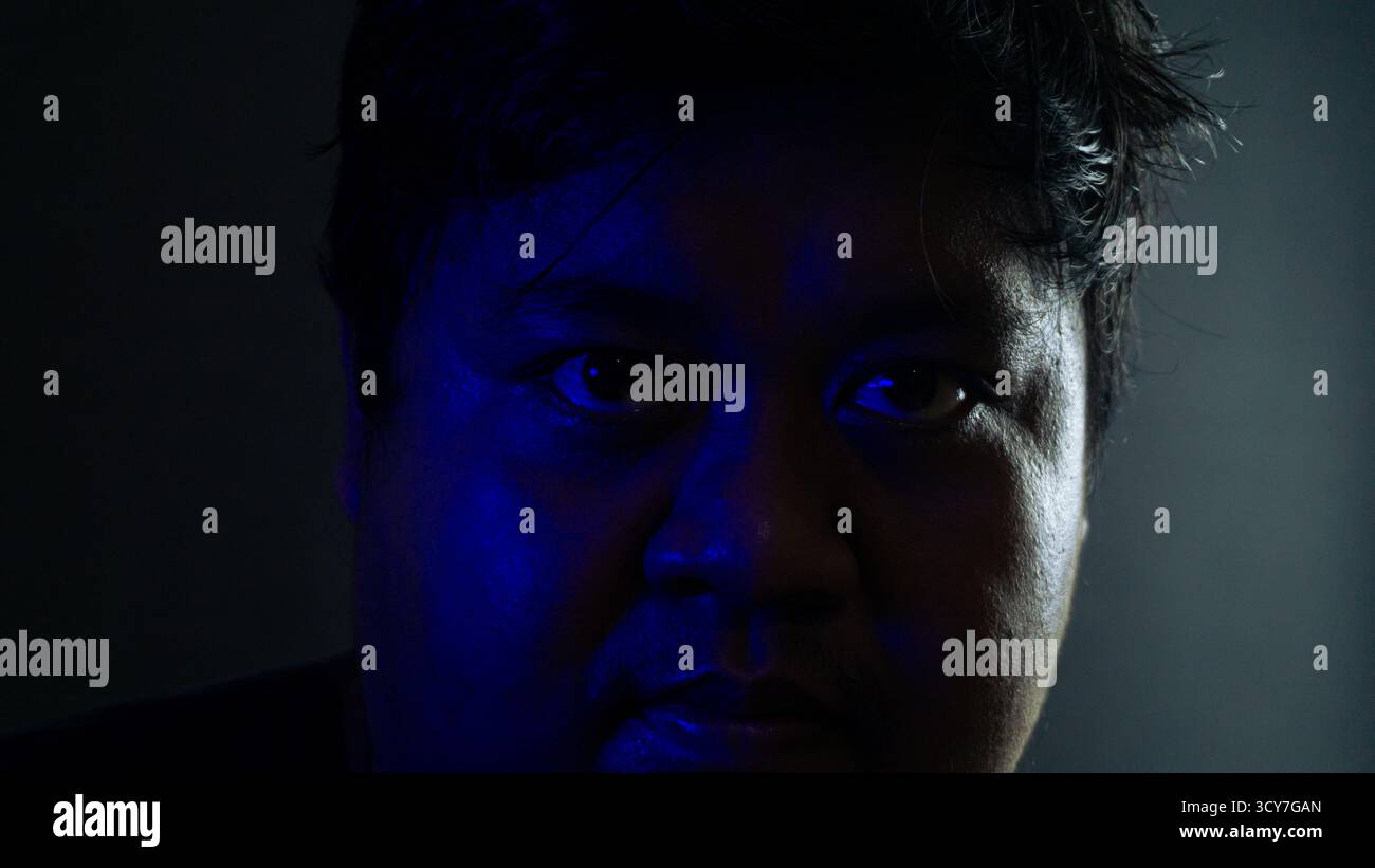 Nahaufnahme männliches Gesicht, teilweise beleuchtet von blauem Umgebungslicht vor schwarzem Hintergrund, geheimnisvoller und schattenhafter Ausdruck, der spannende kinematische M betont Stockfoto