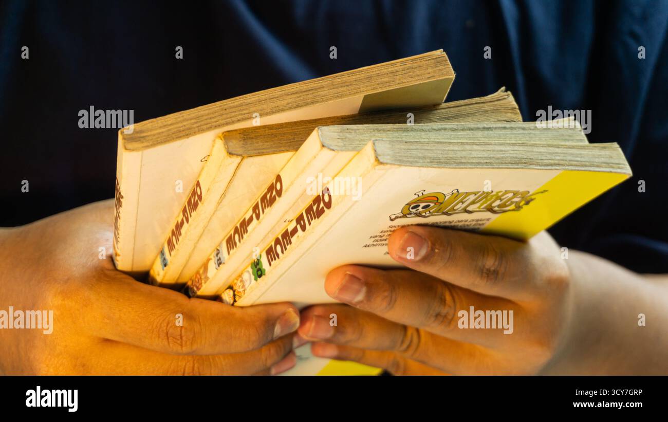 South Kalimantan, Indonesien - 07 21 2025 : ein geschlossener Stapel von Manga-Büchern, die von männlicher Hand unter warmem Umgebungslicht gehalten werden und populäre Comics präsentieren Stockfoto