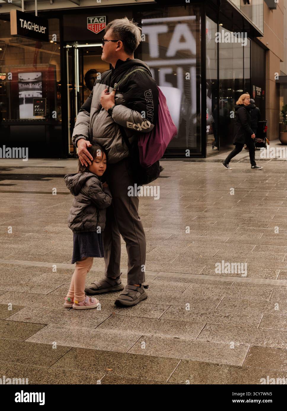 Gedämpftes Foto eines Vaters und einer Tochter, die sich auf einer nassen Straße umarmen; ein zärtlicher Moment des städtischen Schutzes und des Komforts. Stockfoto