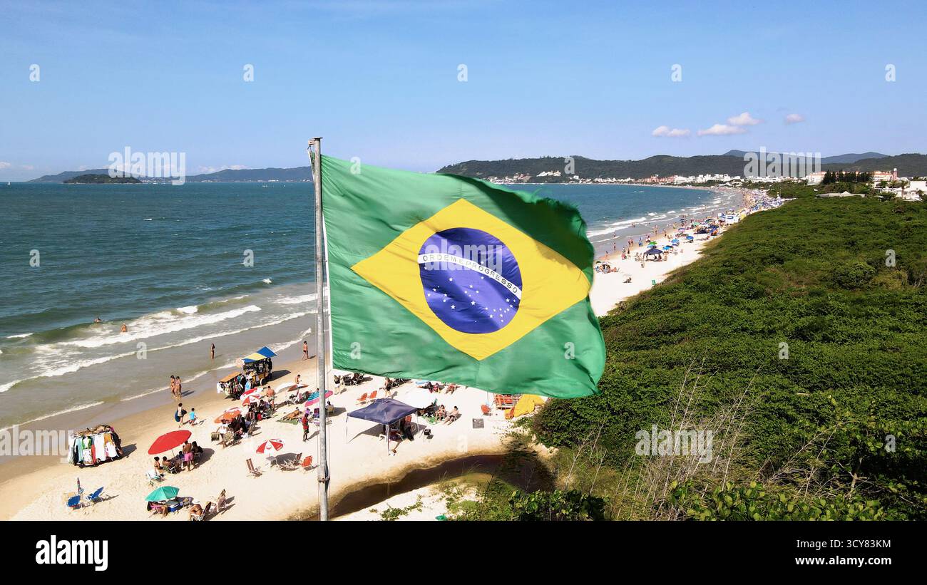 Luftbild der brasilianischen Flagge, die im Wind flattert, am Strand Jurere Internacional in Florianópolis, Santa Catarina, mit dem Strand voller p Stockfoto