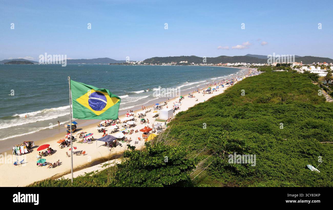 Luftbild der brasilianischen Flagge, die im Wind flattert, am Strand Jurere Internacional in Florianópolis, Santa Catarina, mit dem Strand voller p Stockfoto