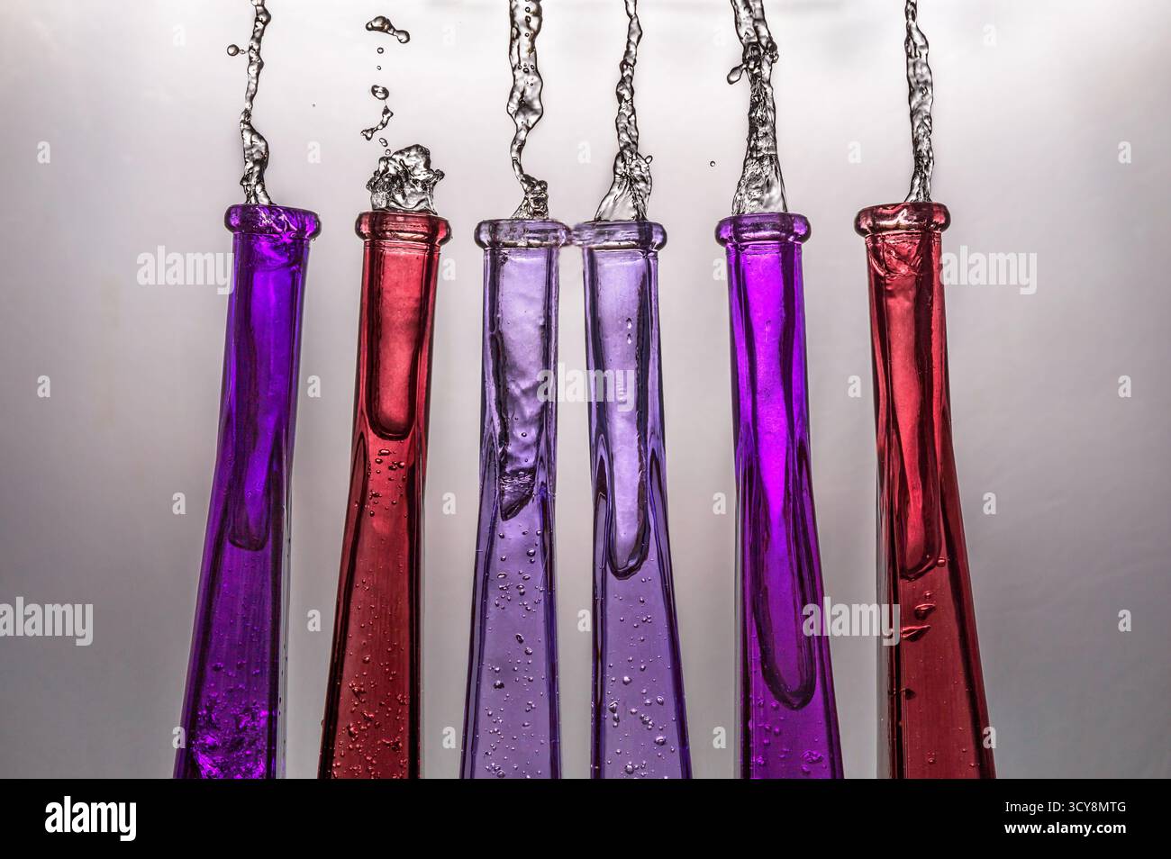 Sechs Glasflaschen werfen farbenfrohe Flüssigkeiten in vertikalen Spritzern aus. Lebhafte Farbtöne deuten auf Cocktails oder starke Spirituosen in dynamischer Bewegung hin. Stockfoto