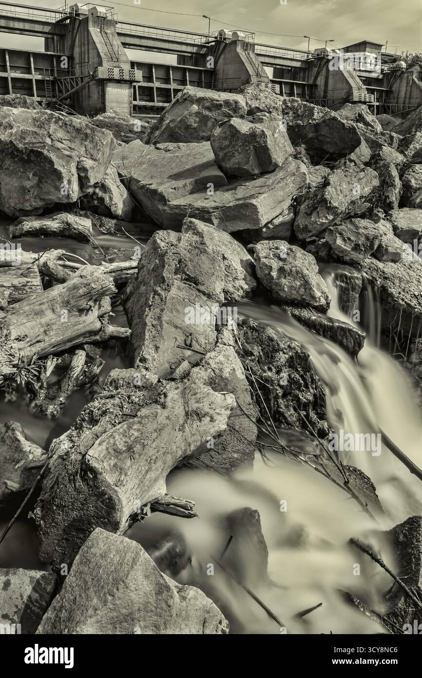 Lange Exposition von Wasser, das über Felsen in der Nähe des rumänischen Damms fließt. Monochrome Töne heben den Kontrast zwischen Natur und Technik hervor. Stockfoto