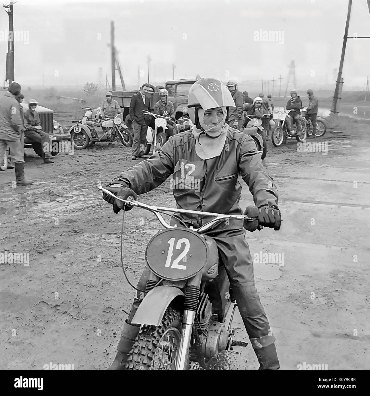 Ein dynamisches Archivfoto aus den 1970er Jahren fängt die düstere Atmosphäre eines Motocross-Rennens in Sloviansk, der ukrainischen SSR, ein. Fahrer mit Vintage-Helmen und Schutzausrüstung sitzen auf ihren leistungsstarken Motorrädern aus der Sowjetzeit (wahrscheinlich CZ- oder Izh-Modelle) auf einer schlammigen Feldpiste, bereit für den Wettkampf. Zuschauer treffen sich in der Nähe, was die Spannung des lokalen Motorsport-Events noch verstärkt. Dieses Bild zeigt die Leidenschaft für Sport und Gemeindeversammlungen in einem friedlichen Donbass vor dem Krieg Stockfoto