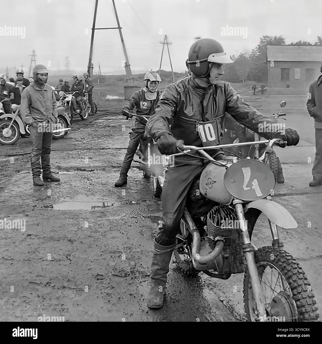Ein dynamisches Archivfoto aus den 1970er Jahren fängt die düstere Atmosphäre eines Motocross-Rennens in Sloviansk, der ukrainischen SSR, ein. Fahrer mit Vintage-Helmen und Schutzausrüstung sitzen auf ihren leistungsstarken Motorrädern aus der Sowjetzeit (wahrscheinlich CZ- oder Izh-Modelle) auf einer schlammigen Feldpiste, bereit für den Wettkampf. Zuschauer treffen sich in der Nähe, was die Spannung des lokalen Motorsport-Events noch verstärkt. Dieses Bild zeigt die Leidenschaft für Sport und Gemeindeversammlungen in einem friedlichen Donbass vor dem Krieg Stockfoto