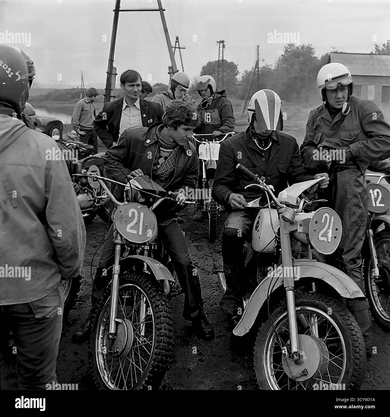 Ein dynamisches Archivfoto aus den 1970er Jahren fängt die düstere Atmosphäre eines Motocross-Rennens in Sloviansk, der ukrainischen SSR, ein. Fahrer mit Vintage-Helmen und Schutzausrüstung sitzen auf ihren leistungsstarken Motorrädern aus der Sowjetzeit (wahrscheinlich CZ- oder Izh-Modelle) auf einer schlammigen Feldpiste, bereit für den Wettkampf. Zuschauer treffen sich in der Nähe, was die Spannung des lokalen Motorsport-Events noch verstärkt. Dieses Bild zeigt die Leidenschaft für Sport und Gemeindeversammlungen in einem friedlichen Donbass vor dem Krieg Stockfoto