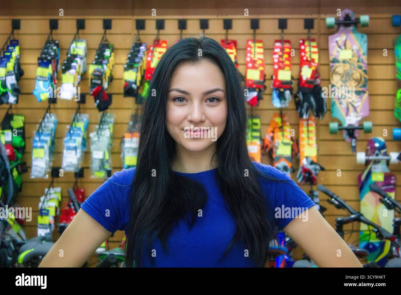 Lächelnde Einzelperson mit langen Haaren in blauem Oberteil in einem Sportgeschäft, in farbenfroher Fahrradausrüstung. Stockfoto