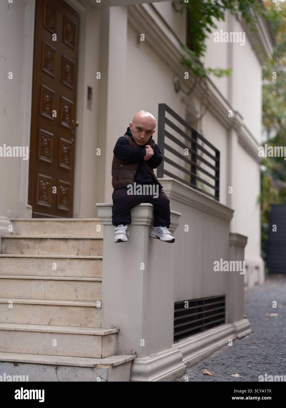 Mann mit Zwergwuchs, der auf einer niedrigen Steinmauer sitzt, Arme an der Brust gefesselt, mit entschlossenem und fokussiertem Ausdruck. Einzigartige Person in URBAN en Stockfoto