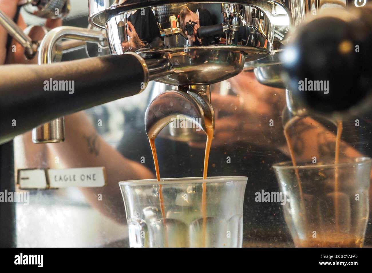 Nahaufnahme eines Espresso, der aus einer verchromten Kaffeemaschine in der Cafeteria Triple Five in Glasbecher gegossen wird, mit sichtbaren Reflexen des Barista. Stockfoto