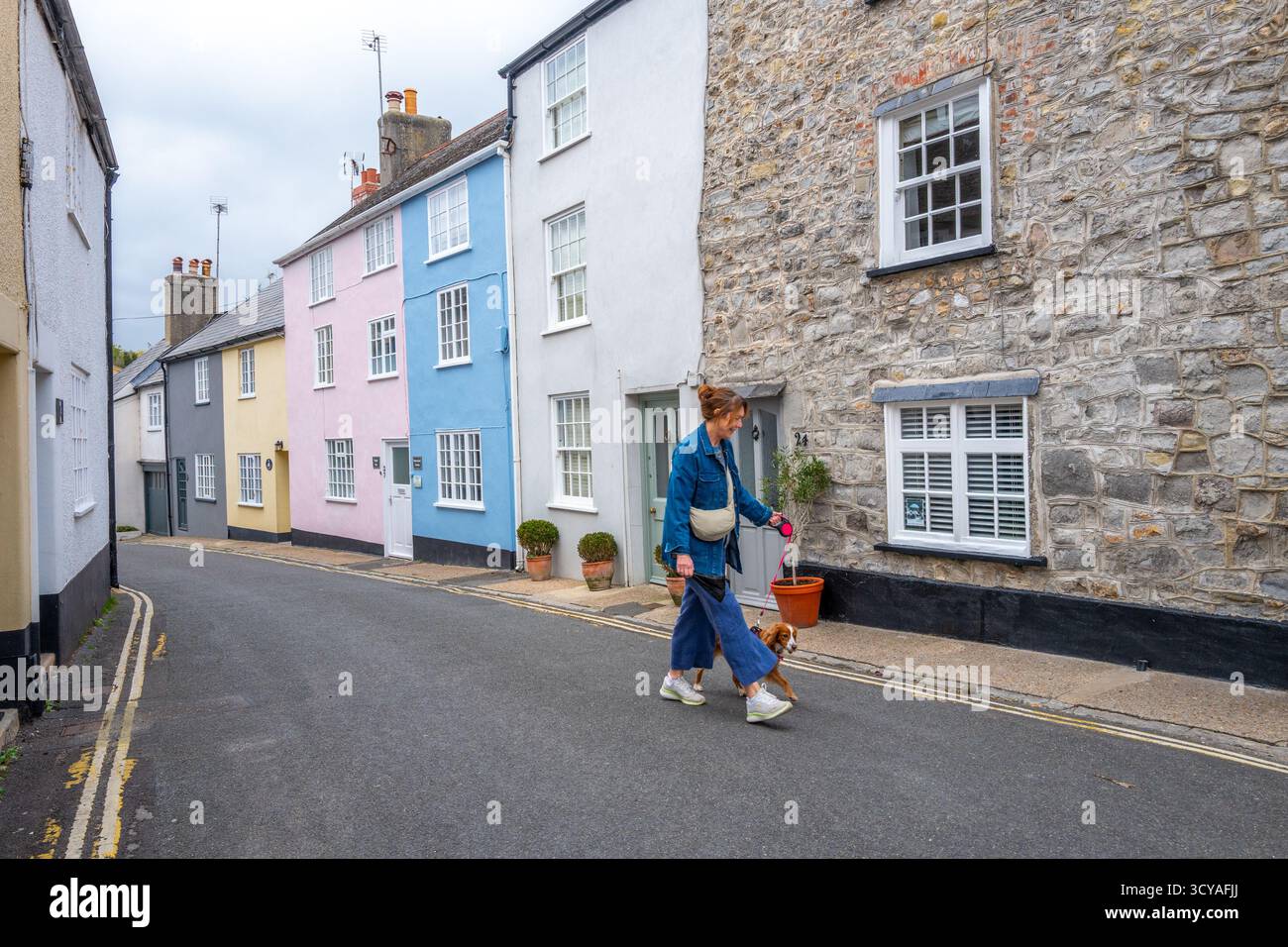 Frau, die ihren Hund an Pastel bemalten Häusern entlang der schmalen Gasse Mill Green in der Küstenstadt Lyme Regis, Dorset, vorbeiführt. Stockfoto