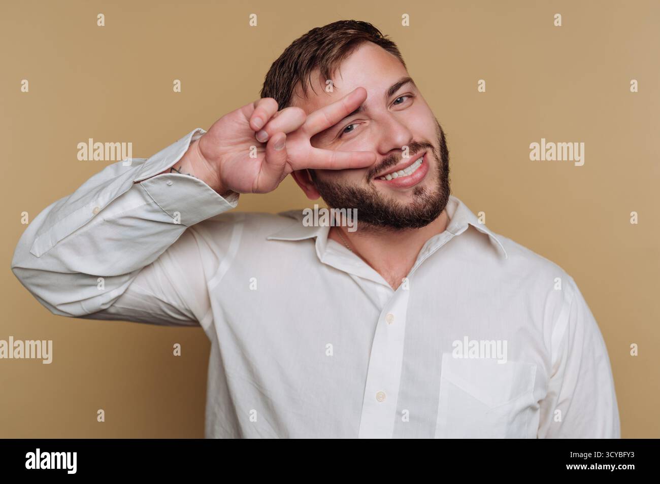 Ein junger Mann in einem weißen Hemd lächelt hell, während er mit einem Friedenszeichen posiert. Der Hintergrund ist ein sanfter, neutraler Ton, der seinen Chee ergänzt Stockfoto