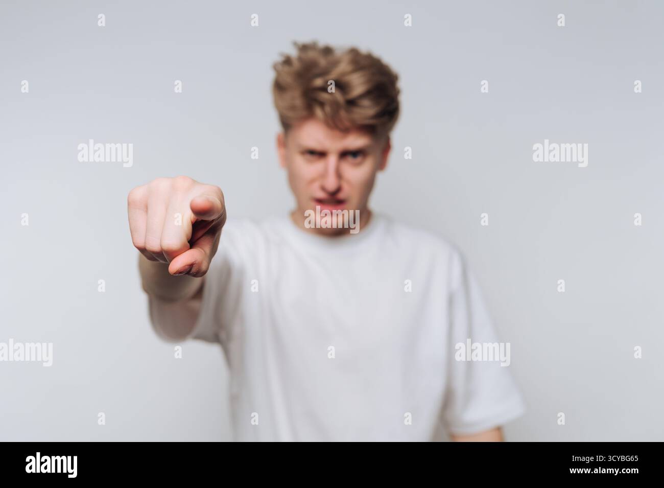 Ein junger Mann steht vor einem schlichten Hintergrund und zeigt mit einem ernsten Ausdruck nach vorne. Seine Geste vermittelt starke Emotionen und schafft ein ansprechendes c Stockfoto Ein junger Mann steht vor einem schlichten Hintergrund und zeigt mit einem ernsten Ausdruck nach vorne. Seine Geste vermittelt starke Emotionen und schafft ein ansprechendes c Stockfoto