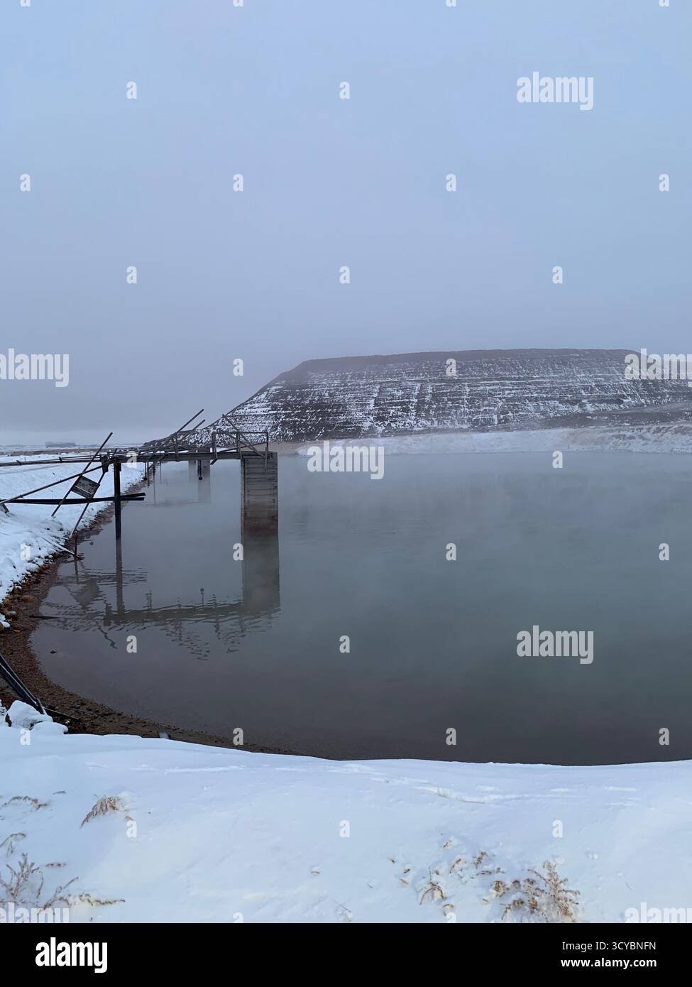 Winterszene an einem abgelegenen Bergbauteich in Kanada mit verwitterter Brücke über dampfendes Wasser, schneebedeckten Böschungen und nebligem Himmel. - Smartphone-aufgenommenes Stockfoto