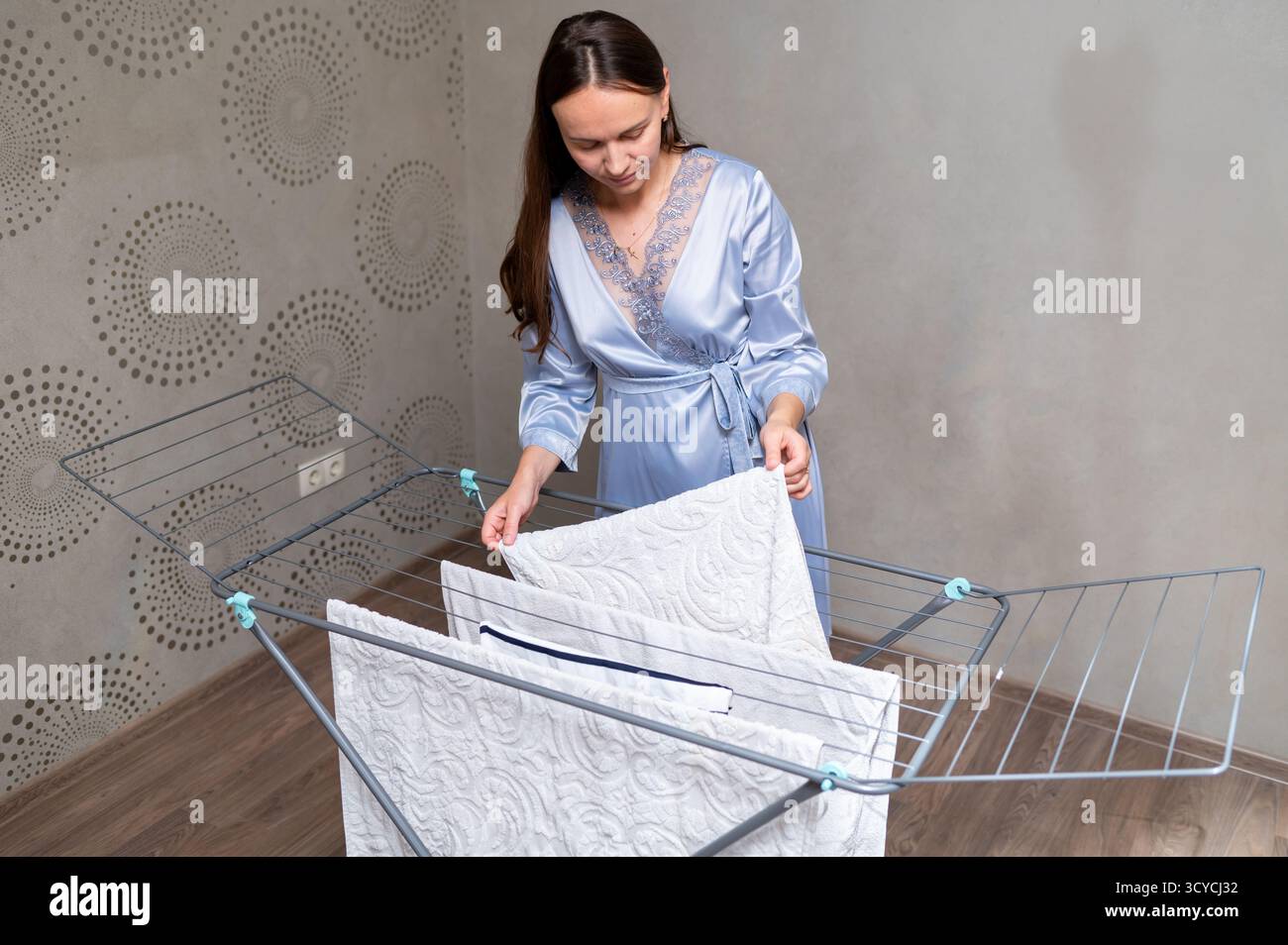 Elegante Hausfrau in Seidenmantel mit lockeren Haaren, die saubere Kleidung auf einem Wäscheständer hängen, Konzept der Hausarbeit und des häuslichen Lebensstils Stockfoto