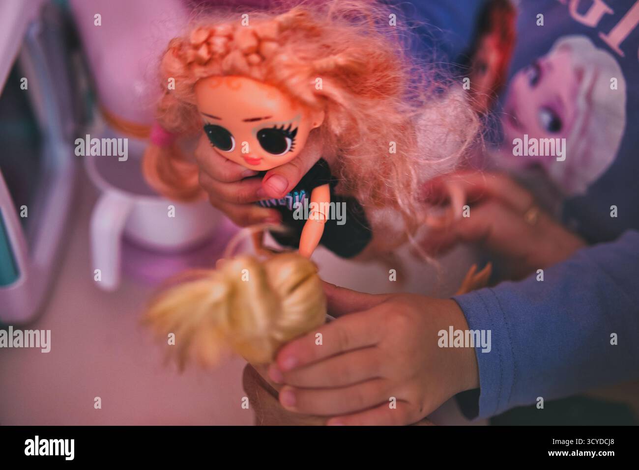 Die Kinderhände halten die unordentlich behaarte LOL Surprise Puppe mit großen Augen gegen das gefrorene Shirt in leuchtendem rosafarbenen Spielzimmer-Setting spielerisch in den Händen Stockfoto