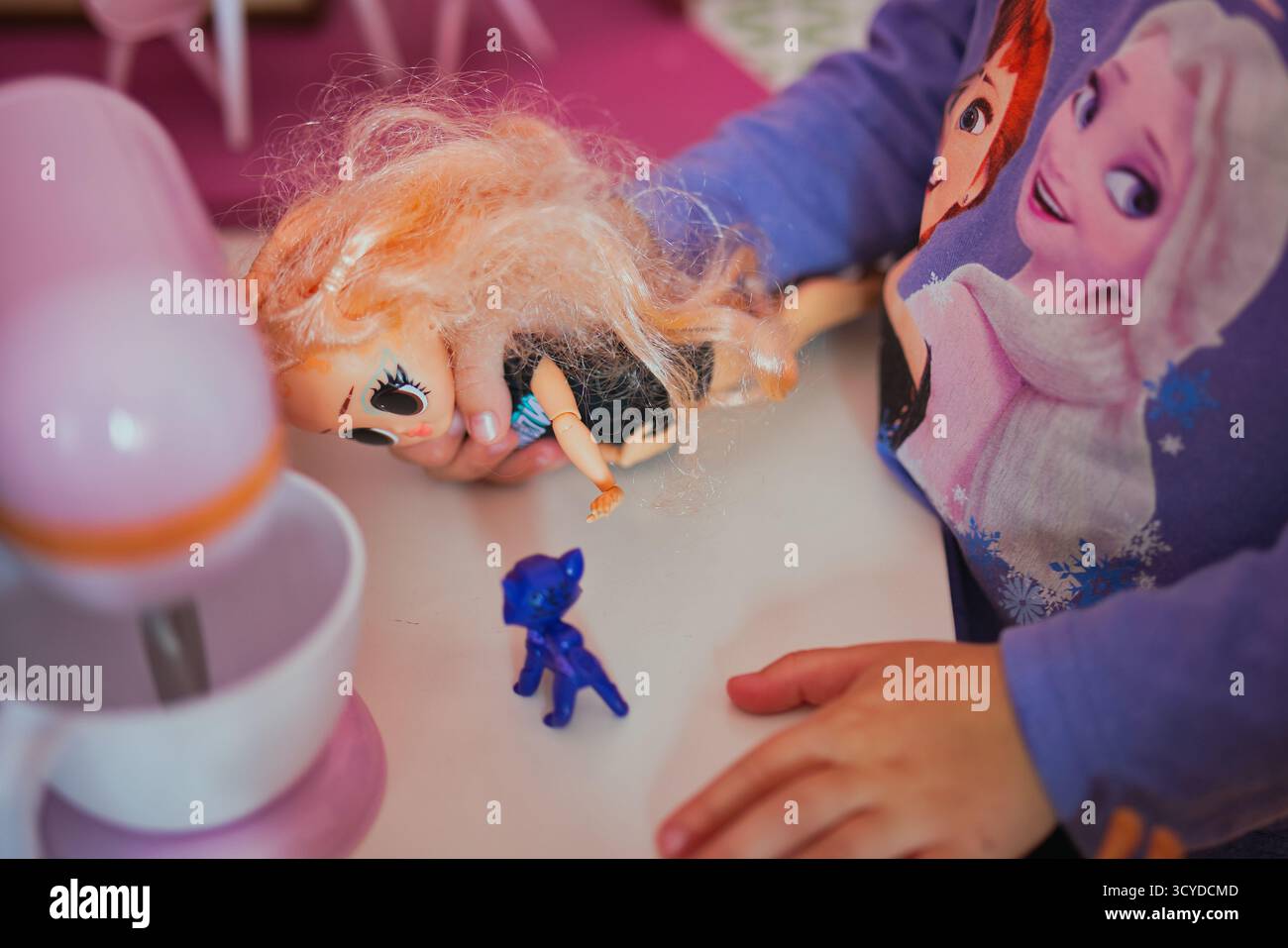 Die Kinderhände halten die unordentlich behaarte LOL Surprise Puppe mit großen Augen gegen das gefrorene Shirt in leuchtendem rosafarbenen Spielzimmer-Setting spielerisch in den Händen Stockfoto