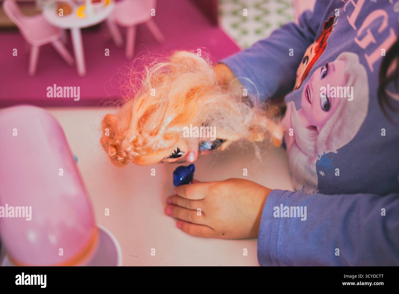 Die Kinderhände halten die unordentlich behaarte LOL Surprise Puppe mit großen Augen gegen das gefrorene Shirt in leuchtendem rosafarbenen Spielzimmer-Setting spielerisch in den Händen Stockfoto