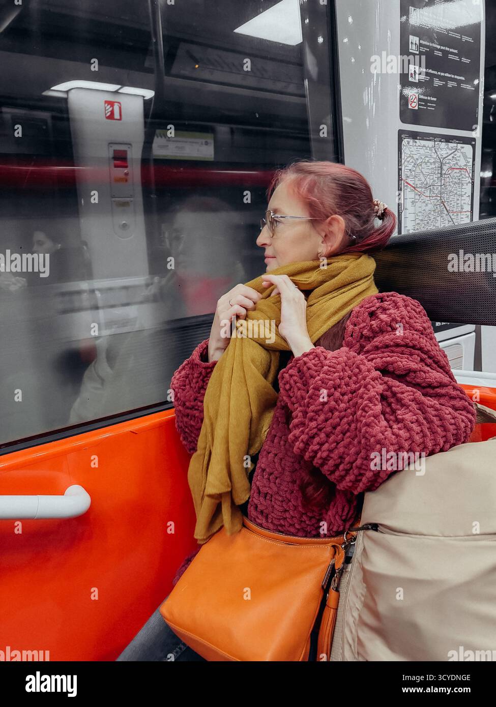 Frau mit orangefarbener Tasche und Senfschal, die in der Straßenbahn sitzt und aus dem Fenster auf unscharfe Reflexionen blickt, trägt einen strukturierten roten Pullover in introspektiver Stimmung Stockfoto