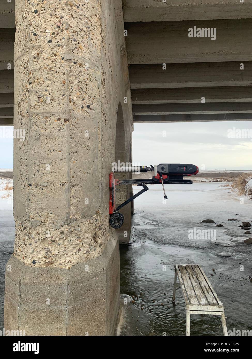 Reparatur von Winterbrücken: Kernbohrer montiert auf verschneiten Betonpier über eiskaltem Fluss, kalte Bauszene. - Smartphone-aufgenommenes Stockfoto